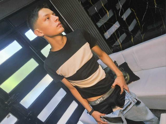 Photograph of SamuJasper, lean and muscular, wearing a black and white striped shirt, pulling down gray pants, exposing a circumcised, semi-erect penis. Black background, white couch, black curtains.