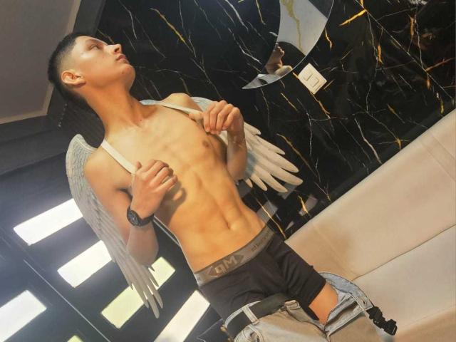 SamuJasper, un hombre musculoso sin camisa con alas grises, está de pie en una habitación tenuemente iluminada, con el pene parcialmente expuesto, pantalones negros. Con la cabeza gacha, mano en el pecho, mirando hacia arriba, pene flácido. Fondo de pared de mármol.