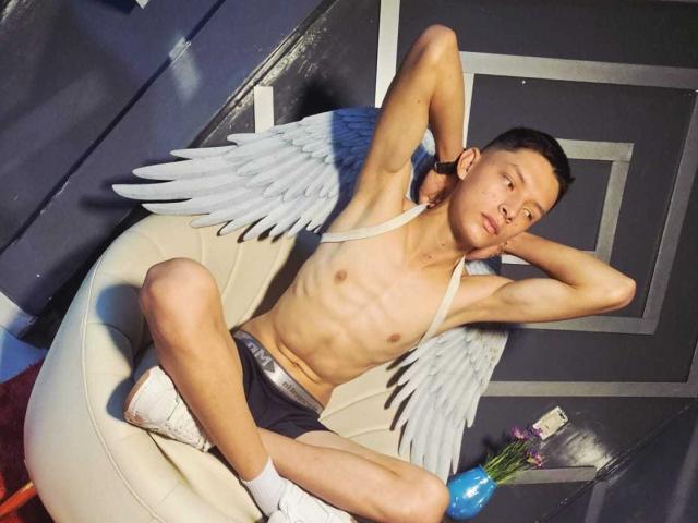 Fotografia de um homem asiático, magro e sem camisa, com asas de anjo brancas, sentado em uma cadeira branca, com as pernas abertas e as mãos atrás. cabeça, shorts pretos, meias brancas, pênis visível, pose sensual.