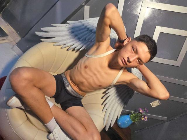 Photographie de SamuJasper, torse nu et musclé, avec des ailes d'ange, allongé sur une chaise crème. Portant un slip noir et des chaussettes blanches, les mains derrière la tête, il montre ses abdominaux bien dessinés. Lumière vive, jeu d'ombres.