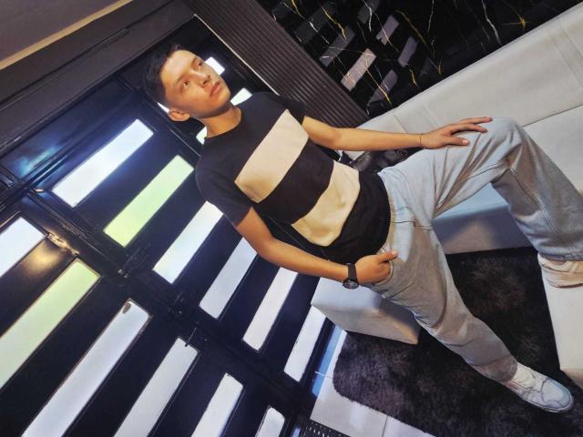 SamuJasper está de pie, con una mano en el bolsillo, apoyado contra una pared blanca. Llevaba una camisa a rayas blancas y negras, pantalones grises claros y zapatillas blancas. Fondo a rayas blancas y negras. Pose casual..