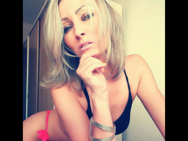 VIPLara, une femme séduisante aux cheveux blonds mi-longs, portant un soutien-gorge noir et des culottes roses, penchée en avant avec sa main près de... sa bouche, regardant séductrice la caméra, portant des bracelets en argent.