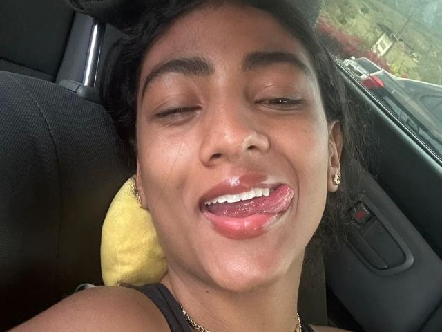 Close-up photo of NatalyBlonder with dark skin, black hair, and shiny lips sticking out tongue. She's wearing a black top and gold earrings. Car seat and yellow pillow in background. Fucking hot.