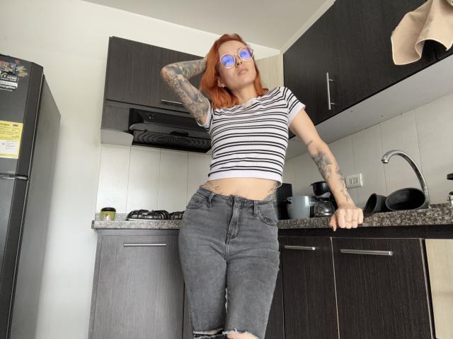 Sarah Rousen steht in einer modernen Küche und lehnt sich mit einer Hand an eine Arbeitsplatte, während die andere Hand auf ihrem Kopf ruht. Sie trägt ein gestreiftes Crop-Top, zerrissene Jeans und blaue, spiegelnde Sonnenbrillen. Sichtbare Tätowierungen an ihren Armen.