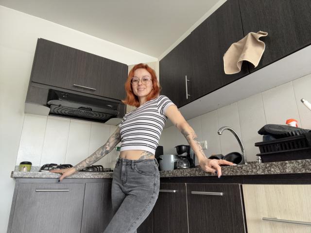 Sarah Rousen se apoya en una encimera de la cocina, vistiendo una camiseta corta a rayas y unos vaqueros ajustados, mostrando tatuajes en su brazo derecho. brazo. Su cabello rojo y su postura segura irradian un atractivo sensual. Se pueden ver objetos de cocina..