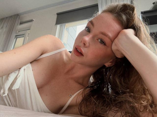 ElliyWilson è sdraiata su un letto, con la testa appoggiata alla mano, e indossa una canottiera bianca a spalline sottili. Ha i capelli castano ondulati, la pelle chiara e un'espressione sensuale e seducente. La luce naturale mette in risalto il suo viso.