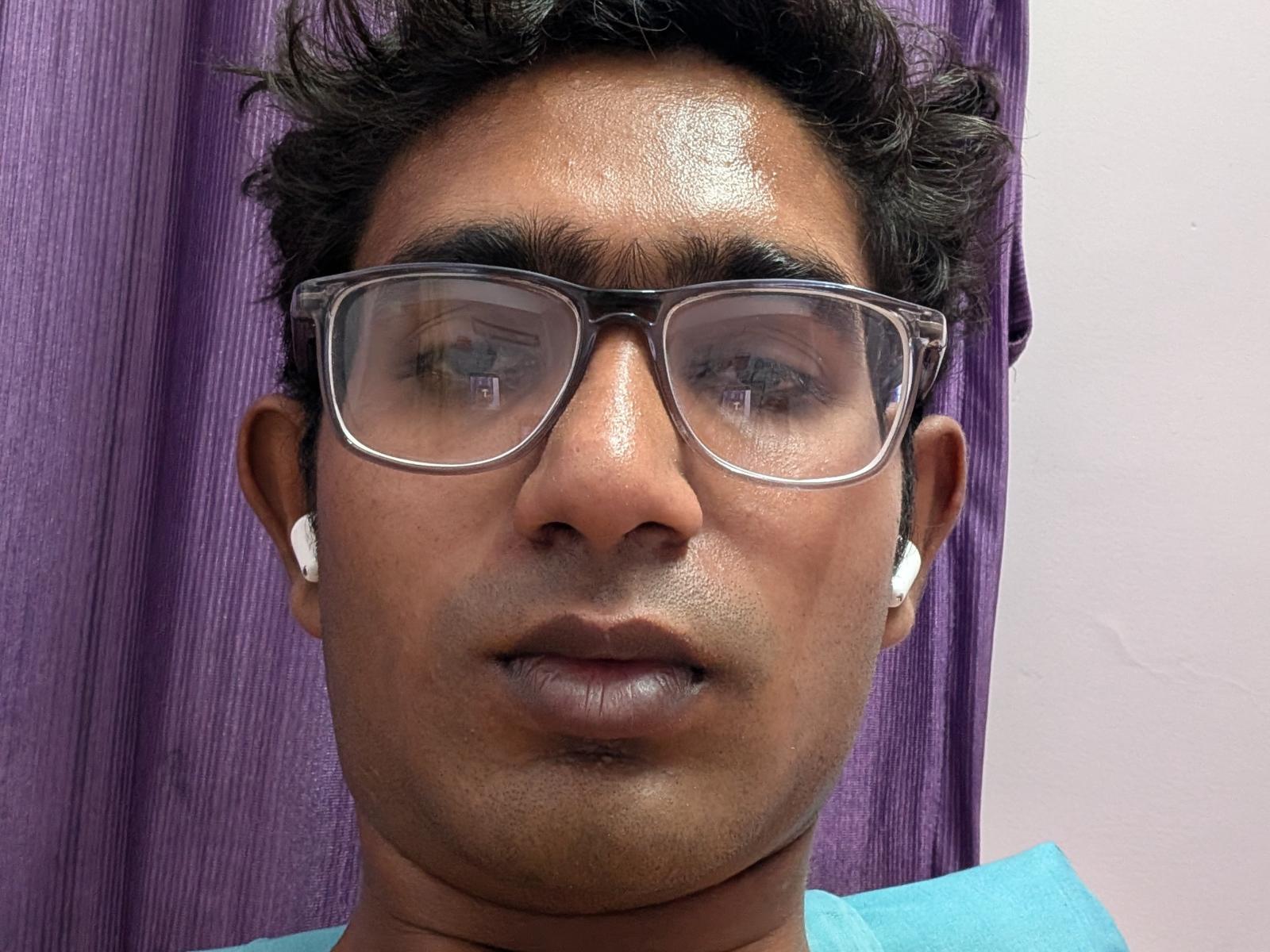 Primer plano de Rubster, con piel de color marrón medio, cabello negro corto y rizado, y gafas transparentes. Está usando auriculares blancos y una camisa azul. Hay una cortina morada en el fondo. Expresión concentrada.