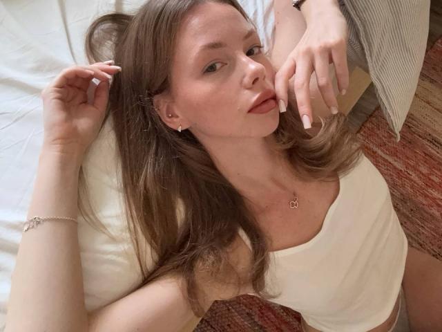 ElliyWilson sdraiata su un letto, con i capelli castani sparsi, indossa una canotta bianca, una mano tocca il viso, l'altra è sulla testa. cuscino a righe, tappeto, braccialetto, tensione sessuale sottile, posa informale.