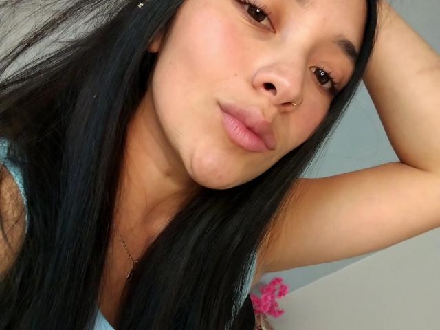 Fotografía de primer plano de ChantalBernadette, una mujer latina con cabello largo y negro, ojos marrones y un piercing en la nariz, que está mirando. Mirando directamente a la cámara con un ligero ceño fruncido, con el brazo levantado. Increíblemente atractivo/a..