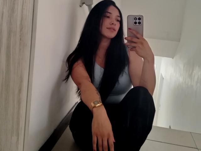 ChantalBernadette, com cabelos longos e pretos, vestindo uma camiseta branca e calças pretas, agachada enquanto tira uma selfie no espelho. com um relógio dourado no pulso esquerdo. Muito atraente, sorrindo de forma sedutora..