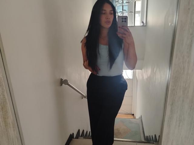 ChantalBernadette prend un selfie devant un miroir dans un couloir étroit. Elle a de longs cheveux noirs, un débardeur blanc et un pantalon noir. Elle tient un téléphone dans sa main droite, et elle a l'air confiante..