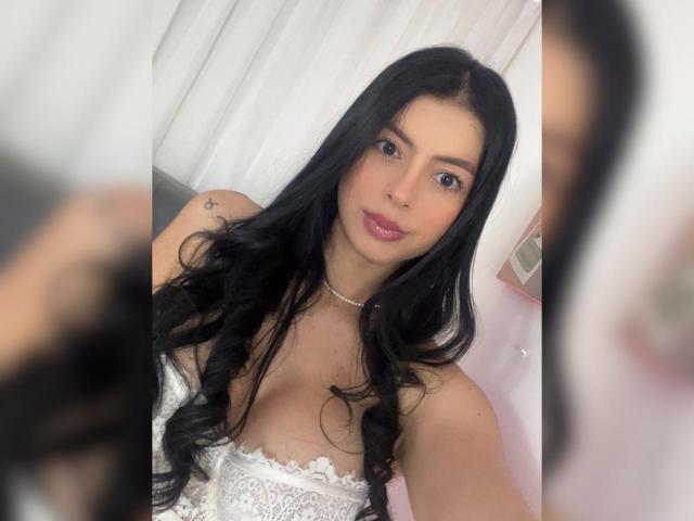 Photograph of IsabellaJames, a woman with long black hair, wearing a white lace strapless top, looking directly at the camera with a slight smile. Blurred figure to the right.