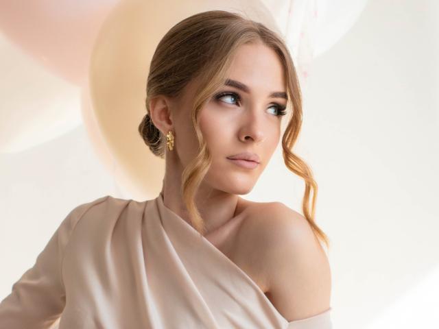 Photograph of EvaDaly, looking over her shoulder with a seductive gaze. She has wavy blonde hair, wearing a silky, off-shoulder cream dress and gold earrings. Her skin is fair, eyes are blue.