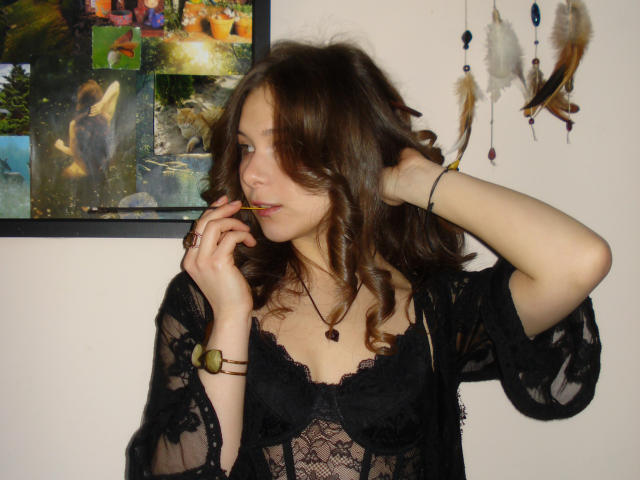 LanaLovely, avec ses cheveux bruns ondulés, porte un haut en dentelle noire, elle mord une sucette, une main dans ses cheveux, une tension sensuelle. Bracelets noirs, collier avec pendentif. Peinture abstraite colorée et attrape-rêves en arrière-plan..