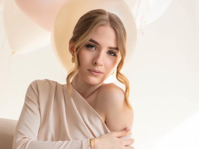 Photographie d'EvaDaly, à la peau claire et aux cheveux bruns tressés de manière lâche, portant un haut blanc à une épaule et des boucles d'oreilles dorées. et un bracelet en or, regardant directement l'appareil photo avec un sourire subtil. Lumière vive en arrière-plan.