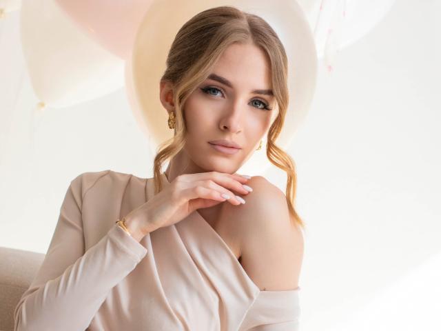 Fotografia di EvaDaly, con la pelle chiara e i capelli castani raccolti in una treccia laterale, che indossa una top beige con una spalla scoperta, con la mano vicino al collo. luce soffusa, espressione leggermente seducente, trucco delicato, orecchini dorati.