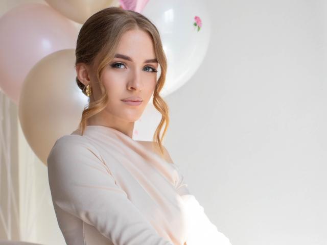 Eva Daly, de piel clara y cabello ondulado castaño, vestida con una blusa blanca de manga larga, está de pie frente a un fondo rosa y blanco. globos, mirando por encima del hombro con una mirada seductora, iluminación brillante..