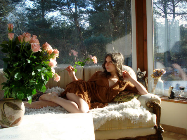 LanaLovely lounges on a sunlit, vintage couch, legs spread, wearing a brown dress, sunlight casting shadows on her face, large pink flowers on the left, window reflecting trees. Fucking relaxed vibe.
