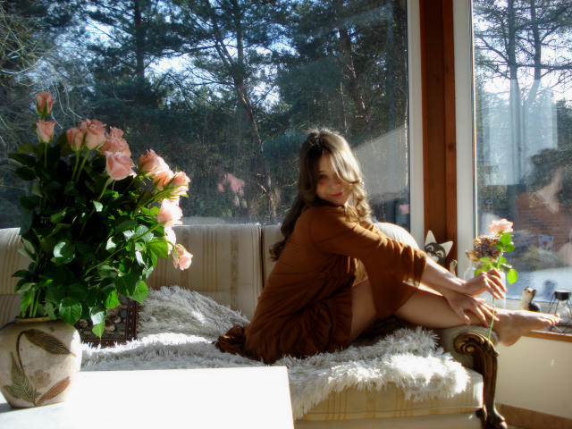 LanaLovely est assise sur un canapé éclairé par le soleil, recouvert de fourrure blanche, les jambes écartées, vêtue d'une robe marron, baignée de lumière solaire. Des roses roses dans un pot sur la gauche, une fenêtre en arrière-plan. Pose séduisante..