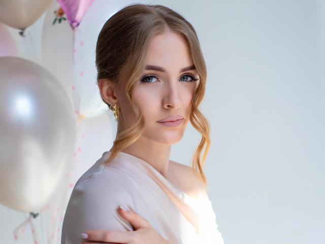 Eva Daly, een vrouw met een lichte huid, blauwe ogen en lichtbruin haar in losse vlechten, draagt een witte top en houdt haar borst vast. staande voor witte ballonnen, helder licht, lichte glimlach.