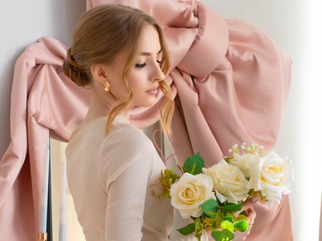 Eva Daly, con i capelli castani raccolti in uno chignon, indossa un abito bianco e tiene un bouquet di rose bianche e gialle, in piedi accanto a tessuto rosa drappeggiato, con uno sguardo di lato e un'espressione seducente..