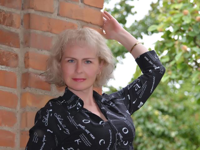 Fotografía de SunnySoul: una mujer rubia, de ojos azules y piel morena, con lápiz labial rojo, una camisa negra con estampado y un brazo levantado. Muro de ladrillos, fondo con hojas verdes, luz suave, expresión ligeramente altanera, pose casual..