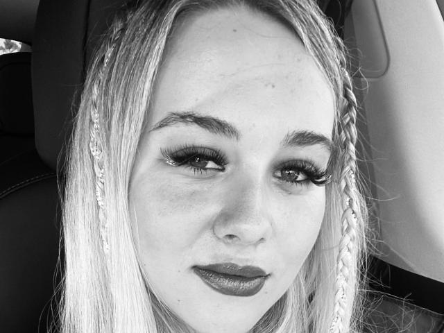 Photo en noir et blanc, en gros plan, d'une femme séduisante, reine du BDSM, avec de longs cheveux blonds, un trait d'eye-liner prononcé et un rouge à lèvres foncé. Son regard est direct, et il exprime une confiance, avec une légère séduction. Contraste élevé.