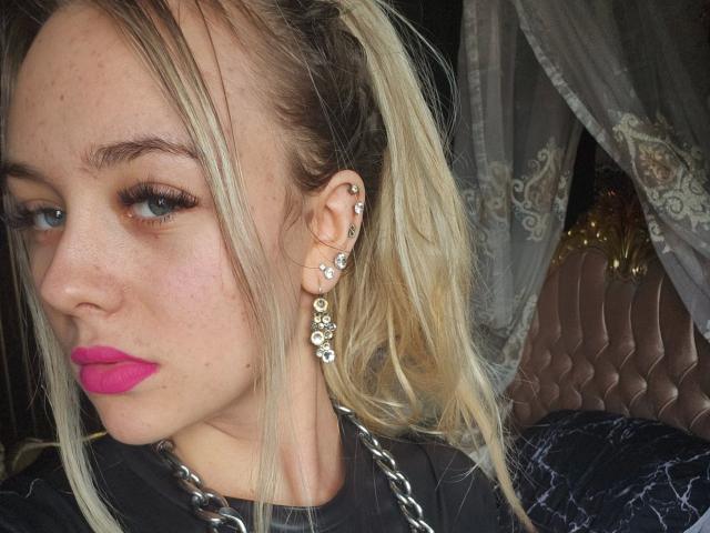 Primer plano de una mujer atractiva, considerada "reina del BDSM", con cabello rubio recogido en una coleta, usando lápiz labial rosa, aretes de plata y una blusa negra. Sus ojos azules miran de lado con una expresión sensual. El fondo muestra cortinas de encaje..