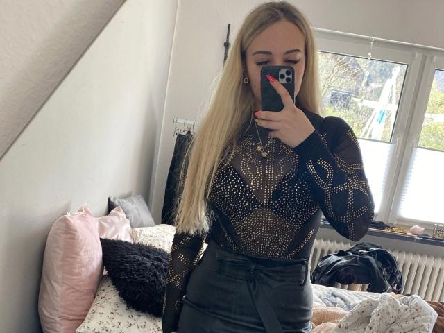 Koningin van BDSM, aantrekkelijk met lang blond haar, draagt een zwarte doorschijnende top en een zwarte broek met hoge taille, en maakt een spiegelzelfportret. Slaapkamer met veel zonlicht, een rommelig bed, een roze kussen en een raam. Verdomd aantrekkelijk..