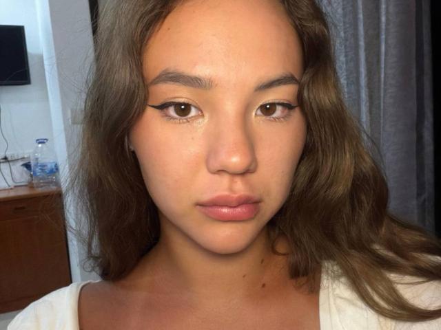 Gros plan de MyrtisTonti, une jeune femme à la peau brun clair, aux lèvres pulpeuses et aux cheveux bruns foncés ondulés. Elle a un trait d'eye-liner ailé, une expression neutre, et porte un haut blanc. Éclairage doux.