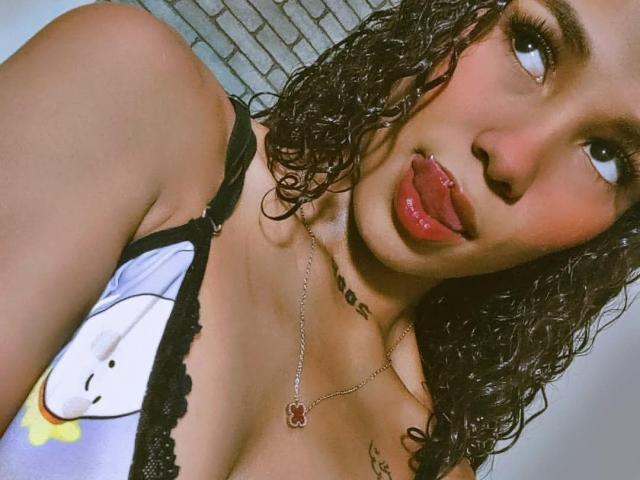 Foto em close-up de LadySeduction, com cabelo cacheado molhado, blusa de renda preta, lábios vermelhos levemente entreabertos, língua para fora, olhos azuis olhando. deitada, colar de ouro com pingente vermelho, expressão sexy e provocante.