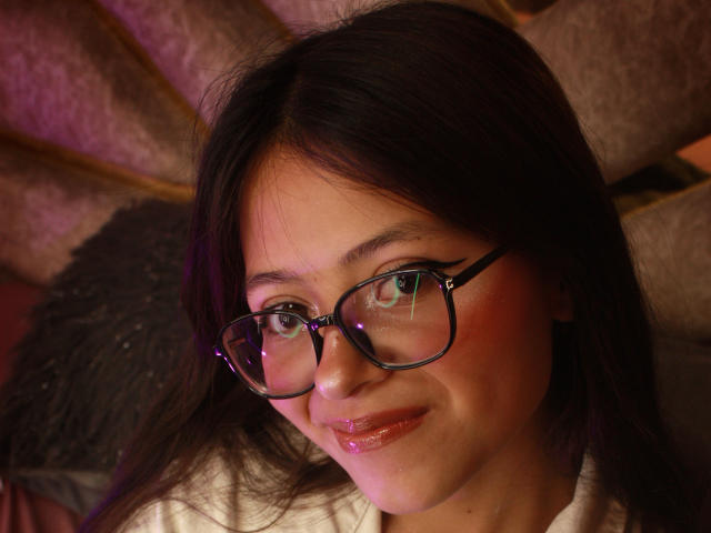 Close-up photo of AnittaRouses, a woman with dark hair, wearing black-rimmed glasses and a slight smile. Her skin has a pinkish glow, and the background is blurred. She's looking directly at the camera.