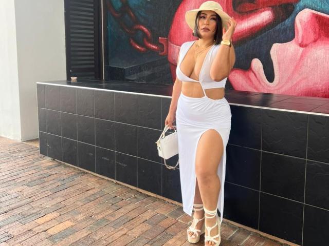 EmaFoxy, de pie frente a una pared de baldosas negras, vestía un vestido blanco de tirantes que dejaba ver mucho, con una abertura alta, tacones blancos y sombrero blanco. Sosteniendo un bolso blanco. Entorno urbano, luminoso..