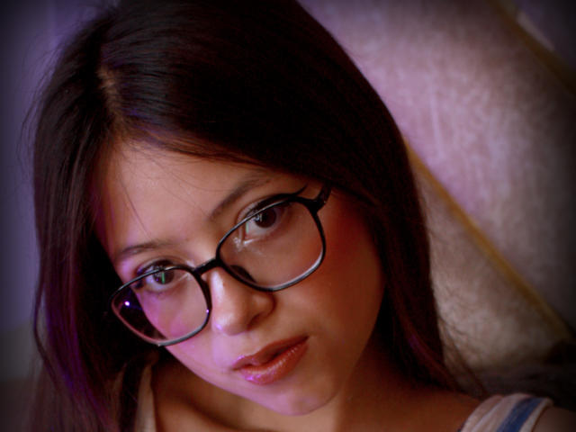 Close-up van AnittaRouses, een jonge vrouw met donkerbruin haar, die een zwarme bril draagt en een subtiele, verleidelijke glimlach. Haar roze lippen en glanzende huid contrasteren met de donkere achtergrond. Heel aantrekkelijk..