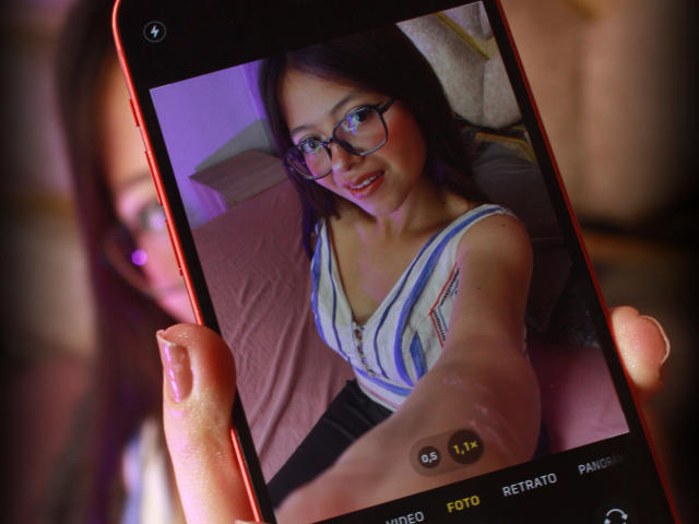 Close-up of AnittaRouses' selfie on a smartphone, holding the phone with her right hand. She's wearing glasses and a striped top, smiling, with visible cleavage and red lipstick. Background blurred.