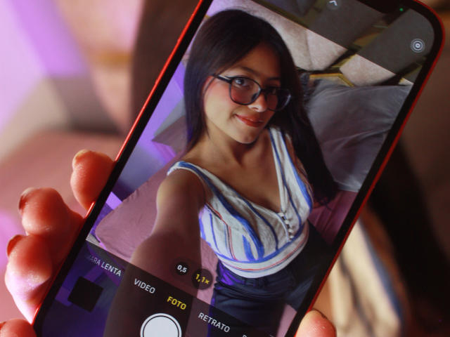 Close-up of a selfie on a smartphone, held by a hand with visible redness. AnittaRouses, with long black hair, glasses, and a blue-striped top, smiles seductively. Soft lighting.