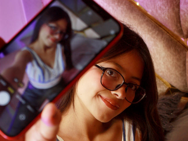 Close-up of AnittaRouses, wearing glasses, smiling, holding a phone with a blurry selfie of herself in a white top, soft lighting, slight focus on her face, intimate, sexy vibe.