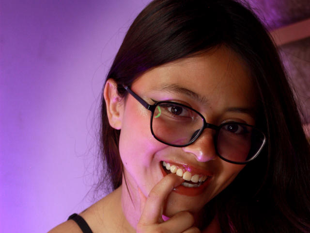 Close-up photo of AnittaRouses with dark brown hair, black-framed glasses, and a mischievous smile. She's biting her finger, eyes slightly squinted, under purple lighting. Fucking cute and sexy.