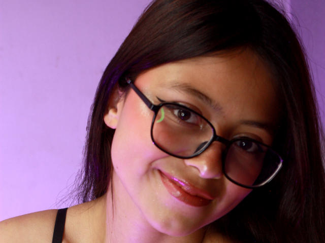 Close-up photo of AnittaRouses with straight black hair, wearing black-framed glasses and a black strap top. She has a slight smile, pink lips, and a slightly flushed face. Soft purple light background.