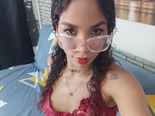 Fotografía de primer plano de LadySeduction, una latina con cabello rizado y mojado, que lleva una blusa roja de encaje, gafas transparentes, lápiz labial rojo y un collar. con colgante, tatuaje en el hombro, mirada intensa, sexy, expresión seductora.