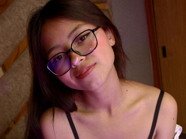 Close-up of AnittaRouses with dark brown hair, wearing black glasses and a black spaghetti strap top. She's looking slightly upwards with a subtle smile, showing light skin and small breasts.