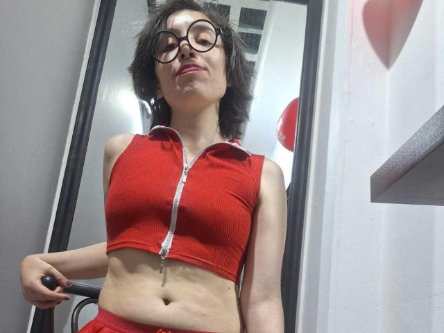Elodie Perrine mit kurzen, zerzausten schwarzen Haaren und Brille, trägt ein rotes, geripptes Crop-Top mit Reißverschluss, das ihre schlanke Taille und kleine Brüste. Der Hintergrund umfasst einen Spiegel und weiße Wände. Verdammt sexy..