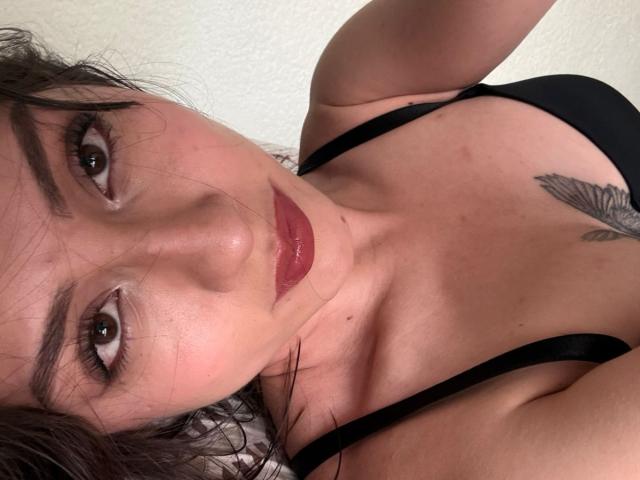 Foto em close-up de Luluberrie, uma mulher de pele clara, com cabelo escuro, batom vermelho e um sutiã preto, com uma tatuagem visível no peito. Ela está olhando diretamente para a câmera com um olhar intenso e sedutor..