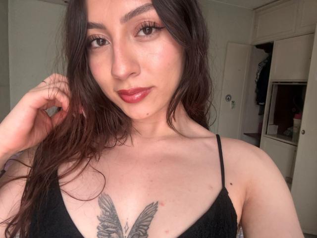 Selfie de perto de Luluberrie, com cabelos longos e castanho-escuros, batom vermelho e uma tatuagem preta de uma borboleta no peito. Usando uma regata preta, com a mão tocando o cabelo. Fundo de quarto..