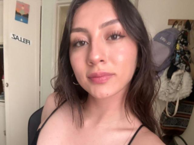 Fotografía de primer plano de Luluberrie, una joven con cabello largo y oscuro, que lleva un top negro con tirantes finos, ligeramente manchado. pintalabios y aros grandes, mirando directamente a la cámara con una sonrisa sutil..