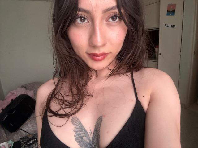Close-up de Luluberrie com cabelo escuro e molhado, pele clara e lábios vermelhos, vestindo uma blusa preta com alças finas que revela tatuagem grande de uma pena preta em seu peito. Olhar confiante, luz natural..
