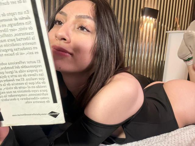 Luluberrie, una donna latina con lunghi capelli neri, è sdraiata a pancia in giù, in lingerie nera, tenendo un libro, con il viso parzialmente nascosta, con un'espressione seducente e guance arrossate.