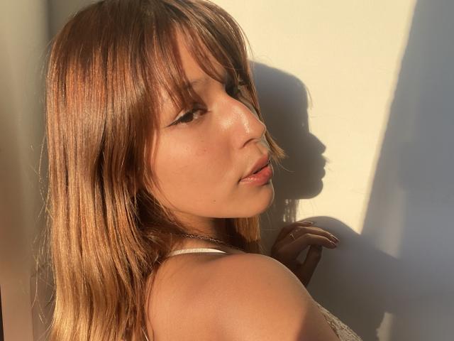 Close-up de KiaraHernandezX, olhando para a esquerda, com cabelo castanho iluminado pelo sol, pele brilhante e expressão sedutora. Sombra à direita, luz vindo da esquerda, realçando sua bochecha e ombro. Incrível, olhando para trás..