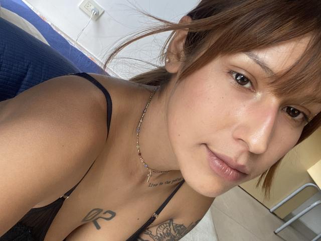 Foto em close-up de KiaraHernandezX, mostrando seu rosto e parte superior do peito. Ela tem cabelo castanho, pele clara e pequenas tatuagens no colarinho e no peito. Usando uma blusa preta, com um leve sorriso. A luz do sol realça sua pele..