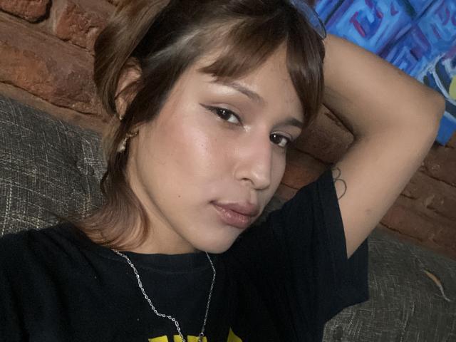 Close-up de KiaraHernandezX, uma mulher asiática de pele clara com cabelo castanho, vestindo uma camiseta preta, com a mão atrás da cabeça. olhar sedutor, leve rubor, ambiente casual, parede de tijolos, tecido azul ao fundo..